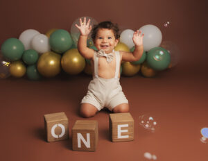 1 year old boy on brown backdrop waving his hands in the air laughing catching bubble during his cake smash photoshoot in whitchurch hampshire