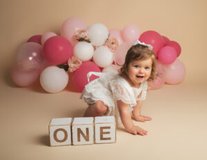 1 year old girl wearing white lace romper on a cream background with pink and white balloon garland