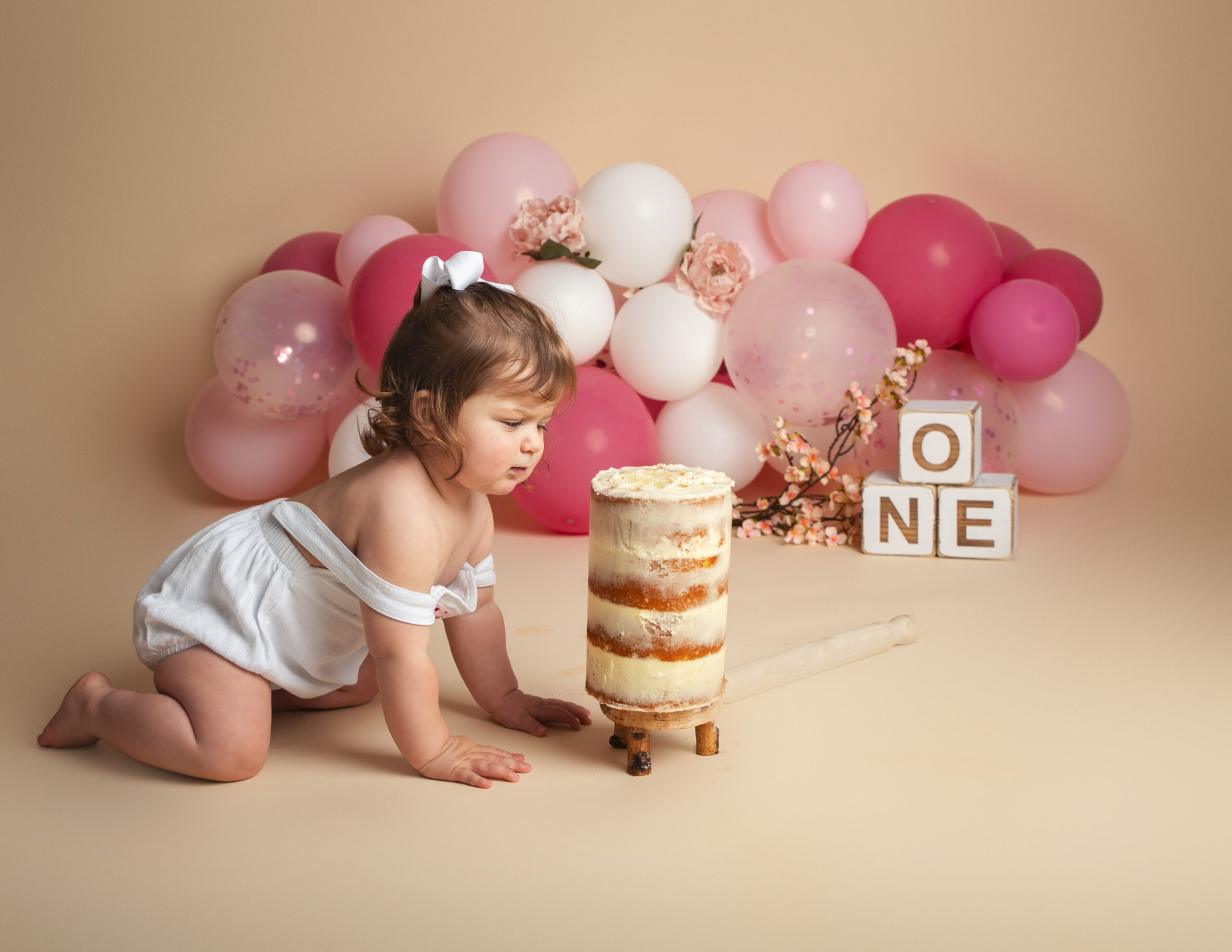 1 year old girl crawling towards a cake ona cream backdrop with pink and white balloons int he background