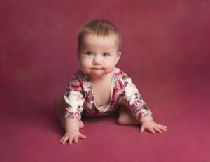 6 month old baby girl on a pink backdrop wearing a white and pink floral vest leaning forward staring into camera