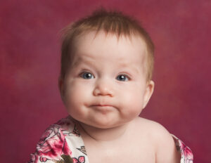 6 month old baby girl frowning into camera on a pink backdrop wearing a floral vest