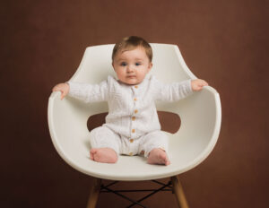 6 month Milestone photo shoot. baby boy sat on large white chair wearing a white knitted romper