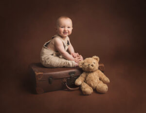 8 month old baby boy sitting ona vintage suitcase witha vintage brown bear smiling at the camera