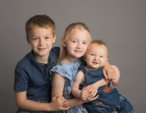 3 children sat ona bench on a grey backdrop smiling at the camera