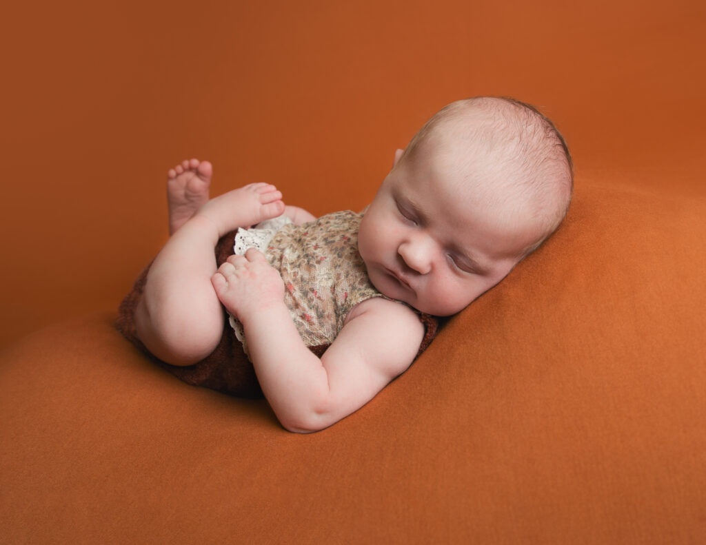 newborn baby girl laying on her back with her legs folded towards her tummy, she is very cutely curled. she is wearing a lace romper and laying on a burnt orange blanket
