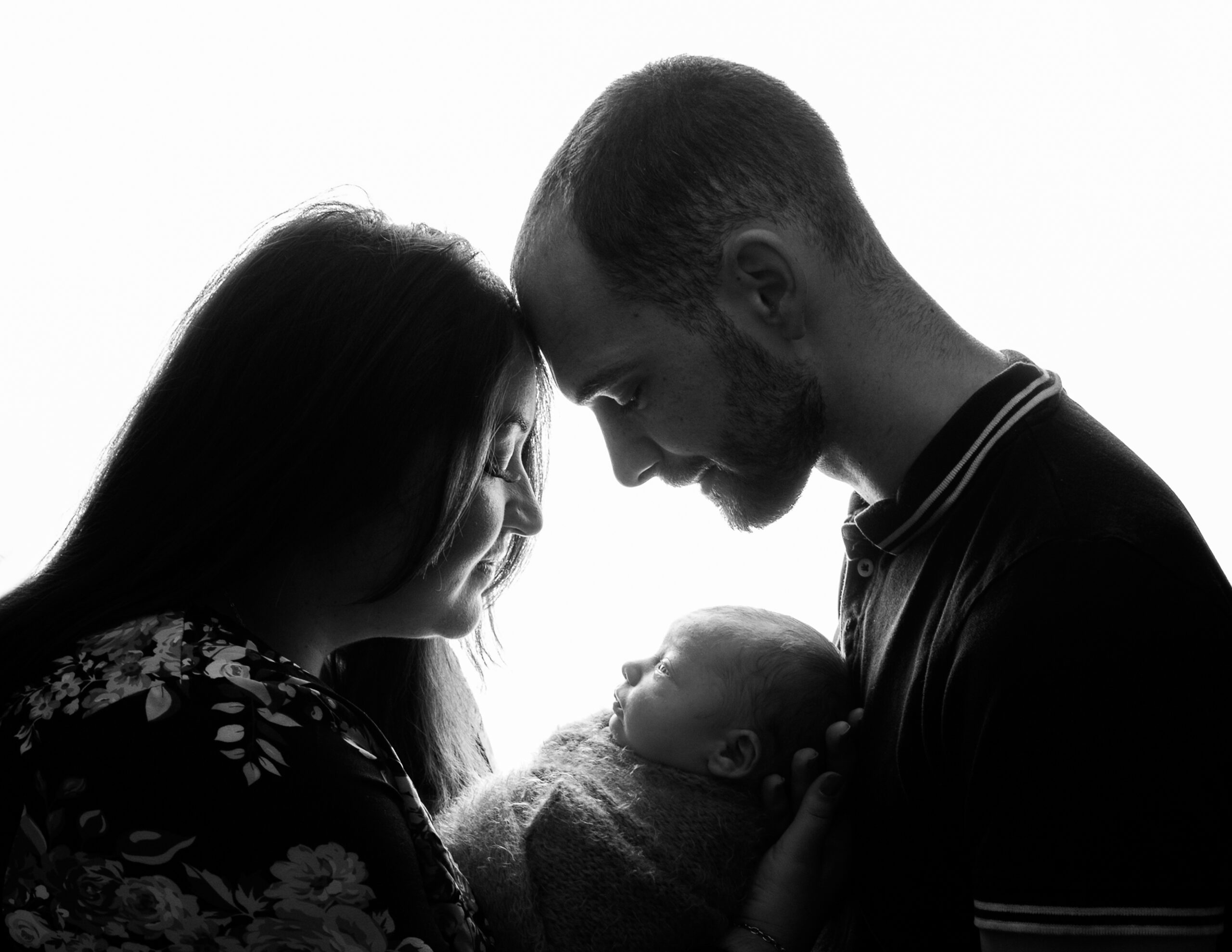 Backlit image of mum, dad and baby with mum and dad looking down on a sleeping baby by Newborn Photographer in Hampshire