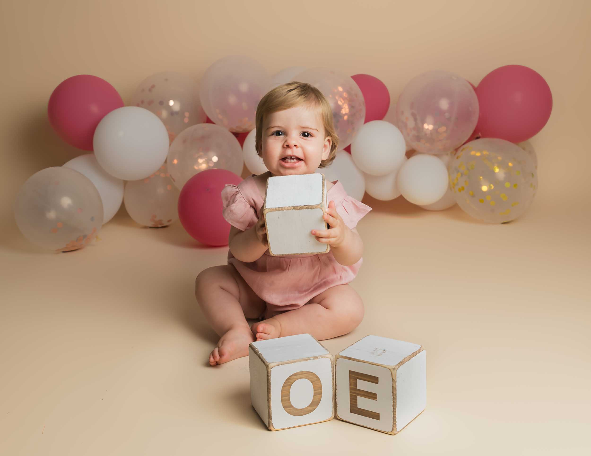 one year old sat on a cream back drop holding a wooden block and smiling at the camera