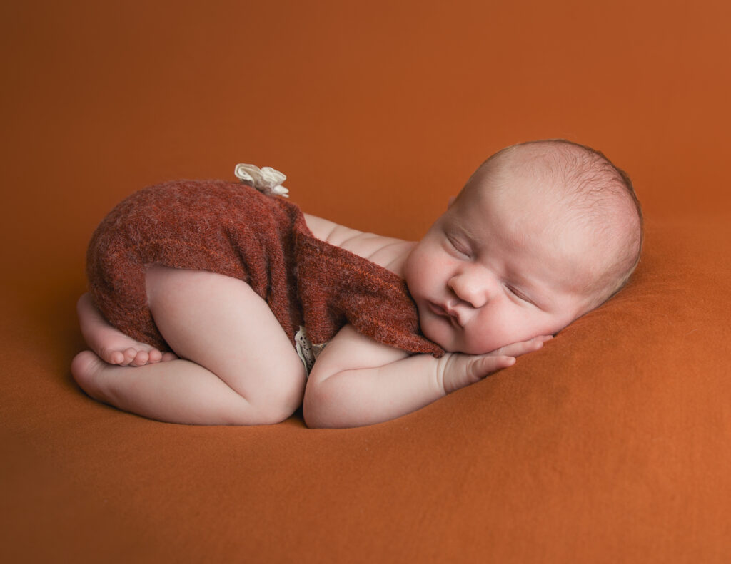 3 week old baby girl in the bum up position on a burnt orange backdrop wearing a burnt orange and lace romper